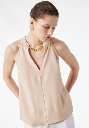 Femme portant un haut beige sans manches à col en V et un collier en chaîne dorée, regardant vers le bas avec les mains derrière le dos.