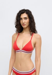 Κόκκινο μπικίνι με τριγωνικό τοπ με λεπτές τιράντες και λευκή και μαύρη ριγέ ταινία στο κάτω μέρος. Ομαλή υφή, μίνιμαλ σχεδιασμός.