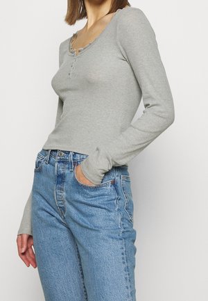 Frau trägt ein eng anliegendes, hellgraues Langarmshirt mit Knopfdetails und hoch taillierte blaue Jeans, eine Hand in der Tasche.