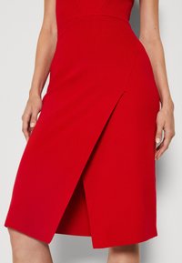 Jupe portefeuille rouge avec un ourlet arrivant au genou, réalisée en tissu texturé, avec des panneaux diagonaux et une fente latérale pour faciliter les mouvements.