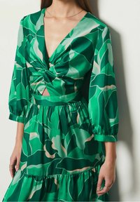 Vestido verde con escote en V, detalle de nudo en la parte frontal, mangas largas y abullonadas, y una falda en capas, con un patrón de hojas en diferentes tonos.
