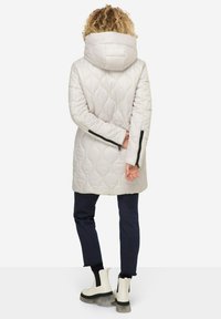 Manteau matelassé beige clair avec capuche, présentant un motif texturé et des accents zippés noirs sur les manches. Porté sur un pantalon bleu foncé et des bottes blanches.