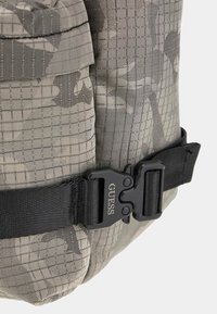 Tarnmuster-Rucksack aus grauem Stoff mit strukturierter Oberfläche; verfügt über einen schwarzen Plastikverschluss mit "GUESS"-Markierung.