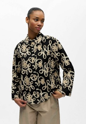 Frau trägt ein schwarzes Hemd mit Knöpfen, verziert mit beigen, bestickten Blumen- und Schlaufenmustern, kombiniert mit hellbeigen Hosen, steht vor einem weißen Hintergrund.