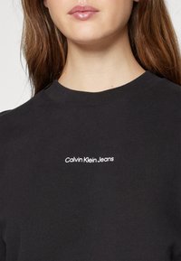 Fekete pulóver bordás gallérral, fehér "Calvin Klein Jeans" logóval a mellrészen. Puha anyag, sima textúrával.