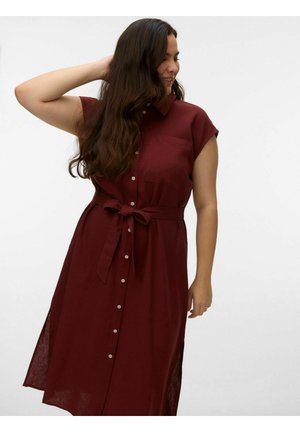 Hosszú sötét hajú nő, aki gombos, ujjatlan, sötétvörös ruhát visel, derekán megkötött övvel, egyszerű fehér háttér előtt áll.