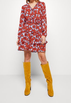 Femme portant une robe bleu à manches longues avec un imprimé floral rouge et des bottes hautes en suède jaune moutarde, debout devant un fond blanc uni.