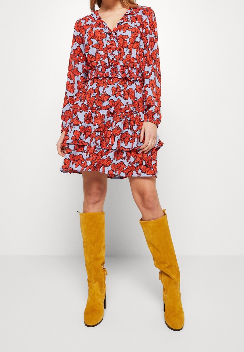 Femme portant une robe bleu à manches longues avec un imprimé floral rouge et des bottes hautes en suède jaune moutarde, debout devant un fond blanc uni.