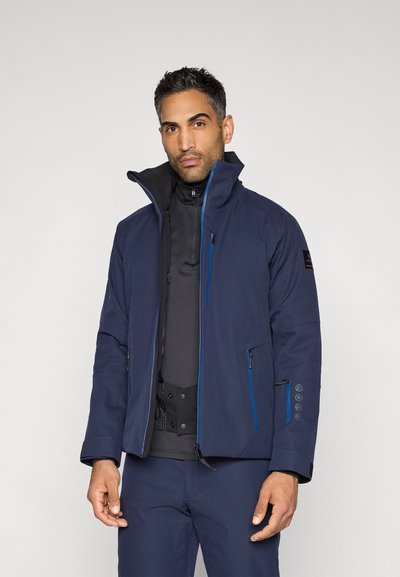 Tamsiai mėlyna neperšlampama striukė su aukštu apykakliu, užtrauktukais užsegamomis kišenėmis, mėlynomis detalėmis ir reguliuojamais rankogaliais. Ant viršaus užsidėta juoda termo palaidinė.