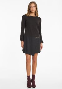 Schwarz langärmliges Kleid mit einem gestreiften asymmetrischen Saum, das mit einem goldenen Akzentdetail und gerafften Ärmeln versehen ist. Textilstruktur und taillierte Passform.