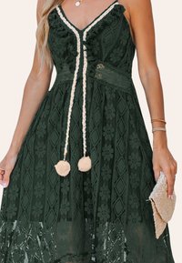 Donkergroene jurk met bloemenkant patroon, V-hals, franjes, gevlochten strikjes en twee pompons. Model houdt een geweven clutch vast.