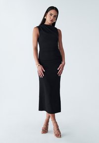 Abito midi senza maniche di colore nero con texture arricciata, scollo rotondo e silhouette aderente. Indossato con tacchi dorati con cinturini.