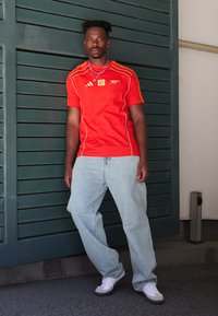 Rode Adidas-shirt met gele details, lichtblauwe baggy jeans en witte sneakers. Staande tegen een donkergroene geslepen achtergrond.