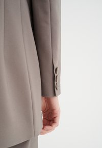 Blazer grigio chiaro su misura con una texture morbida, dotato di uno spacco singolo, tre bottoni sui polsini e una silhouette aderente.