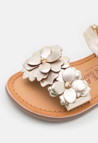 Metalické zlaté sandály s širokým popruhem zdobeným třemi vrstvenými květinovými akcenty, umístěné na hnědé kožené stélce. Design s plochou podrážkou.