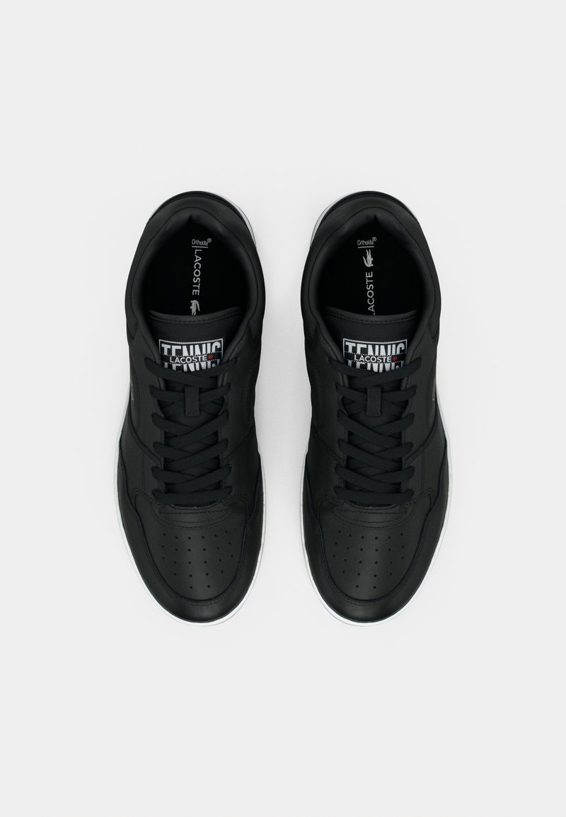 Baskets noires à lacets avec détails perforés, tige en cuir synthétique lisse, col rembourré et semelle extérieure en caoutchouc blanc. Logo Lacoste visible.