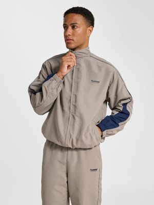 Mann richtet den Kragen einer beige-blauen hummel Trainingsjacke, steht mit einer Hand in der Tasche vor einfachem Hintergrund.