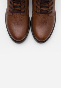 Brązowe skórzane buty o teksturowanym wykończeniu, okrągłym nosku i czarnych sznurowadłach. Posiadają czarną podeszwę z przeszywanym brzegiem.