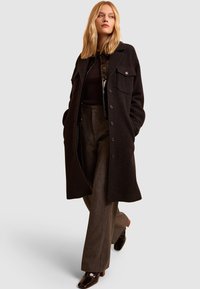 Abrigo largo negro de tejido texturizado, con dos bolsillos en el pecho y cierre de botones, llevado sobre una parte superior oscura y pantalones de pierna ancha grises. Zapatos negros de alto brillo.