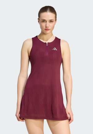 Mujer con vestido deportivo sin mangas color granate con cremallera y logo en el pecho, de pie mirando hacia adelante frente a un fondo liso.