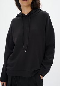 Femme vêtue d'un sweat à capuche noir ample avec cordons, debout devant un fond clair uni, avec une main dans la poche.
