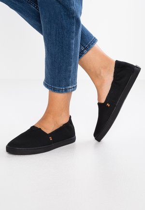 Pieds portant des chaussures slip-on noires en toile avec un petit logo orange, associés à un jean bleu court sur un fond de sol blanc.
