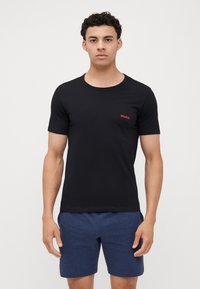 T-shirt noir à col rond en coton lisse, marqué d'un petit logo "HUGO" rouge sur la poitrine. Le modèle porte un short bleu marine.