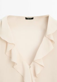 Blusa beige pallido con scollatura arricciata e chiusura a bottoni. Realizzata in tessuto leggero, presenta una texture morbida e un drappeggio delicato.
