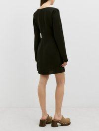 Zwarte jurk met lange mouwen van gestructureerde stof, getailleerd silhouette, ronde halslijn en uitsnijding aan de achterkant, gecombineerd met bruine hoge klompen.