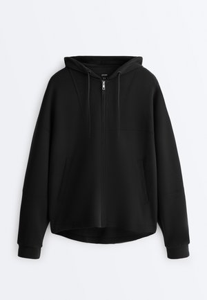 Femme portant un sweat à capuche noir et un pantalon, tenant un sac de sport marron, debout de dos sur un fond blanc.