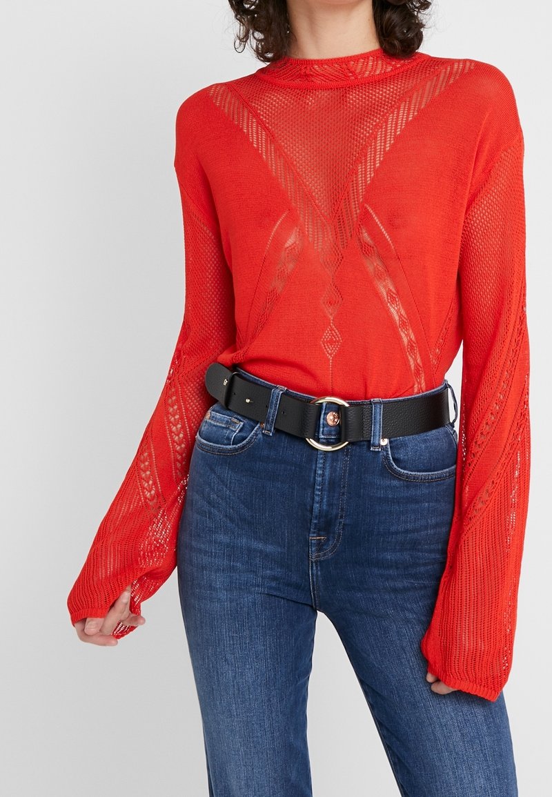 Pull en tricot rouge avec un motif texturé et des manches longues, associé à un jean bleu foncé et une ceinture noire avec une boucle ronde.