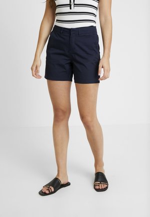 Kvinde iført marineblå shorts, sorte slip-on sandaler og en hvid ærmeløs top med sorte vandrette striber.