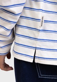 White long-sleeve shirt with blue horizontal stripes, featuring side slits and a fabric label reading "REDEFINED REBEL" near the hem.