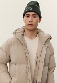 Νέος άνδρας που φοράει ένα μπεζ μπουφάν puffer πάνω από ένα ανοιχτόχρωμο πουλόβερ και ένα σκουρό πράσινο πλεκτό σκουφάκι με λευκά κεντημένα γράμματα.