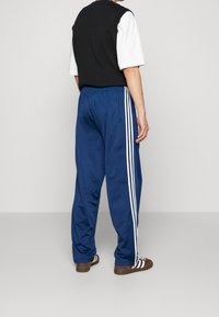 Pantalones deportivos azules con franjas laterales blancas, cinturilla elástica y una textura suave y lisa, combinados con un chaleco negro sobre una camisa blanca.