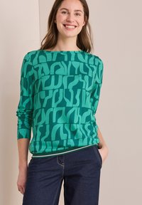 Grön långärmad topp med ett djärvt abstrakt mönster, rundad halsringning och mjuk tyg. Kombinerad med mörka jeans, vilket ger en avslappnad look.