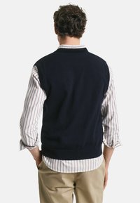 Marine gebreide vest met een gladde textuur, ronde hals en geribbelde onderzoom, gedragen over een wit- en roodgestreept shirt.