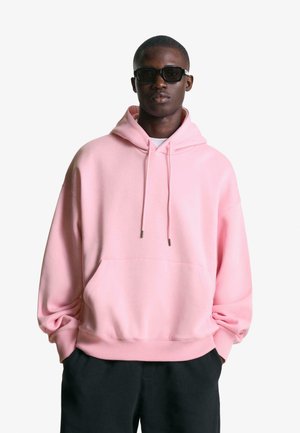Pink oversized hoodie with a drawstring hood, front pocket, and textured fabric. Worn with dark pants, featuring a minimalistic design.