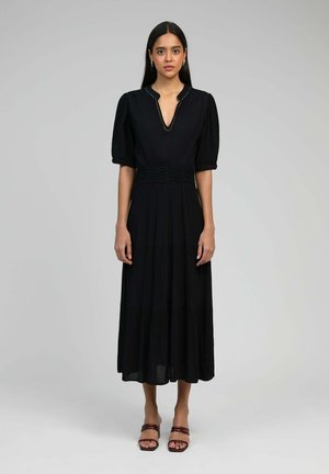 Robe maxi noire avec des manches bouffantes, décolleté en V, taille froncée et jupe à volants. Tissu lisse avec une texture subtile. Le modèle porte des sandales bordeaux.