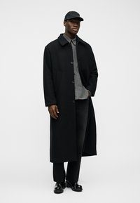 Mann trägt langen schwarzen Mantel über grauem Hemd, schwarze Hose, schwarze Mütze und schwarze Schuhe, steht vor einem schlichten weißen Hintergrund.