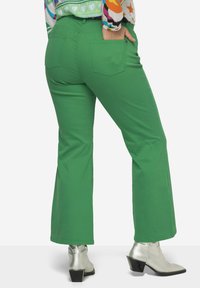 Pantalones acampanados verdes de mezcla de algodón con dos bolsillos traseros. Llevados con botas de tobillo plateadas con tacón cuadrado.