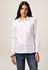 Camicia bianca con bottoni, maniche lunghe e colletto classico, realizzata in tessuto di cotone liscio. Indossata con jeans blu con dettagli decorativi.