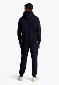 Tumši zils sporta džemperis un nūjotāja komplekts no mīksta auduma ar kapuci un ribveida manšetēm, kopā ar baltām sporta kurpēm. Skats no aizmugures, kas atklāj brīvu piegriezumu.