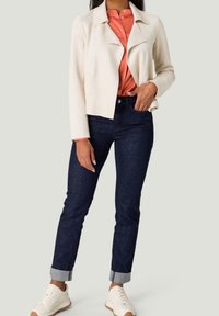 Veste en similicuir crème avec col, associée à un chemisier corail, un jean en denim foncé et des baskets blanches avec des accents tan.