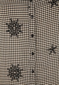 Chemise à carreaux noir et blanc ornée de motifs de flocons de neige brodés en noir, avec des boutons noirs sur le devant et un tissu doux et texturé.