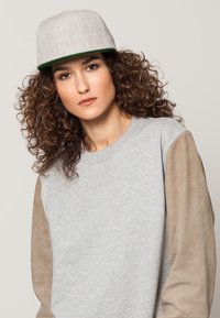 Grijze sweatshirt met beige leren mouwen. Grijze pet met een groene klep. Model met krullend haar kijkt recht vooruit. Eenvoudige achtergrond.