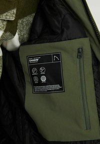 Poche intérieure d'une veste verte avec une étiquette noire indiquant la marque Chasin et des icônes pour les caractéristiques déperlantes, coupe-vent, séchage rapide et maintien de la chaleur.