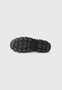 Suola di scarpa nera con un motivo testurizzato e tacchi spessi, caratterizzata da un logo a forma di cuore al centro. Lo sfondo è bianco uniforme.