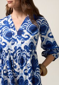 Sininen ja valkoinen kuviollinen mekko, jossa on syvä V-pääntie, kerätty vyötärö ja kolme neljännestä hihaa. Mekossa on kukkakuosi ja pehmeä kangas.