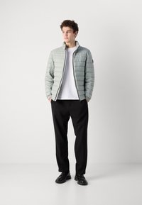 Light green puffer jacket with quilted design, zip front, and high collar. Worn over a white shirt, paired with black trousers and shoes.